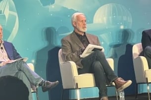 Three caucasian men dressed casually are seated on a stage engaging with the audience.